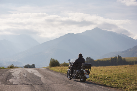 Back view of motorcycler in black leather jacket riding cruiser bike along road on blurred copy space background of beautiful foggy mountain range and blue cloudy sky on bright sunny summer day.の写真素材