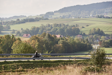 Motorcyclist in helmet black leather clothing riding bike along sharp turn of empty road on bright summer day on misty background of rural landscape and distant green hills.の写真素材