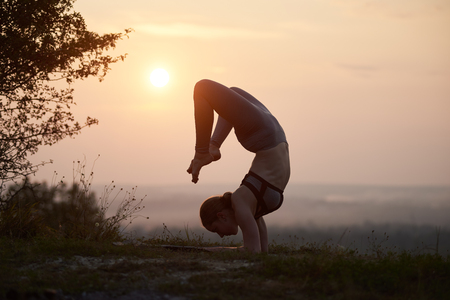 Silhouette of strong flexible barefooted girl in training outfit doing difficult complicated yoga exercises outdoors on background of light pink summer evening sky. Fitness and beauty concept.の写真素材