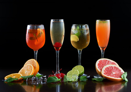 Composition with four different shapes crystal cocktail glasses with cold drinks, orange, lime and grapefruit slices, melting ice cubes, mint leaves and red cherries on black copy space background.の写真素材