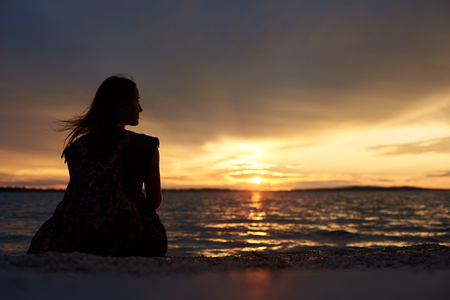 Back view silhouette of tourist woman with backpack sitting alone on seashore at water edge, enjoying beautiful view of sunset on dark evening sky background. Tourism and vacations concept.の写真素材