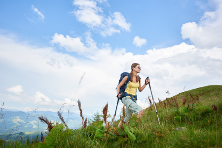 Attractive woman tourist hiking mountain trail, walking on grassy hill, wearing backpack and sunglasses, using trekking sticks, enjoying summer day. Outdoor activity, lifestyle conceptの写真素材