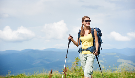 Attractive happy woman tourist hiking in Carpathian mountain trail, walking on grassy hill, wearing backpack, sunglasses, using trekking sticks, enjoying summer day. Outdoor activity, tourism conceptの写真素材