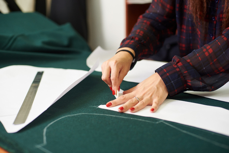 Tailor, designer workplace in atelier. Woman hands making white chalk markings of paper patterns on green fabric copy space surface before cutting details for future jacket or costume.の写真素材