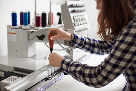 Young woman operator making adjustments in modern computerized embroidery machine using screwdriver on white background. Industrial embroidery, commercial production and high quality results concept.の写真素材