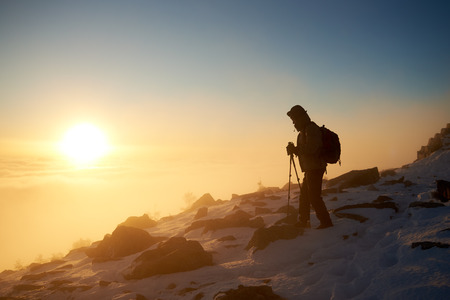 Silhouette of tourist hiker with backpack and trekking poles on rocky snowy mountain steep slope on background of foggy valley filled with white puffy clouds, raising sun and blue sky at sunrise.の写真素材
