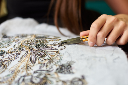 Close-up of female worker hand with special sharp scissors cutting away short loose ends of threads from white fabric detail with machine embroidery of black and yellow floral pattern.の写真素材