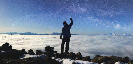 Back view of tourist hiker man with raised arm standing on rocky hill on copy space background of beautiful foggy valley filled with white clouds, snowy mountain tops on horizon and blue starry sky.の写真素材