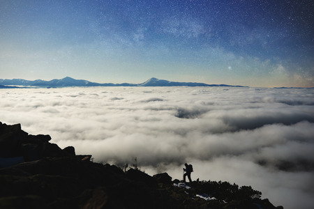 Small silhouette of tourist hiker with backpack and trekking poles climbing steep rocky mountain slope on background of blue starry sky, foggy valley filled with white clouds and snowy mountain tops.の写真素材