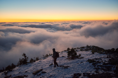 Tourist with backpack hiking on rocky snowy mountain top on background of valley covered with white puffy clouds stretching to foggy horizon under bright morning sky with light orange glow at sunrise.の写真素材