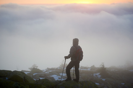 Silhouette of tourist hiker with backpack and trekking poles on mountain big rocks enjoying beautiful view of clouds down below in valley and bright yellow orange dramatic morning sky at sunrise.の写真素材