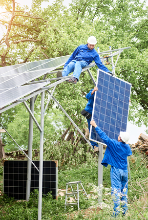 Installing of stand-alone solar photo voltaic panel system. Workers in hard-hats and blue overall lifting the solar module on metal platform. Alternative energy and professional construction concept.の写真素材
