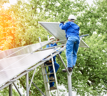 Installing of stand-alone solar photo voltaic panel system. Workers in hard-hats and blue overall lifting the solar module on metal platform. Alternative energy and professional construction concept.の写真素材