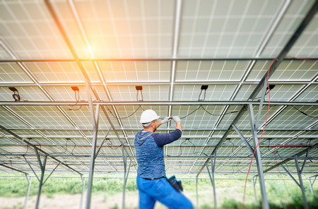 Young electrician in hard hat and rubber gloves wiring solar photo voltaic modules inside the new installed panel system on bright sunny day. Alternative energy and profitable investment concept.の写真素材