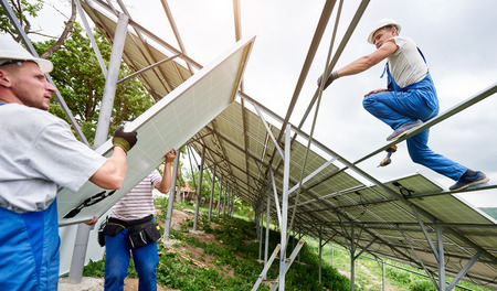 Installing of solar photo voltaic panel system. Three technicians lifting heavy solar module on high platform. Investment in alternative energy, money saving and professional construction concept.の写真素材