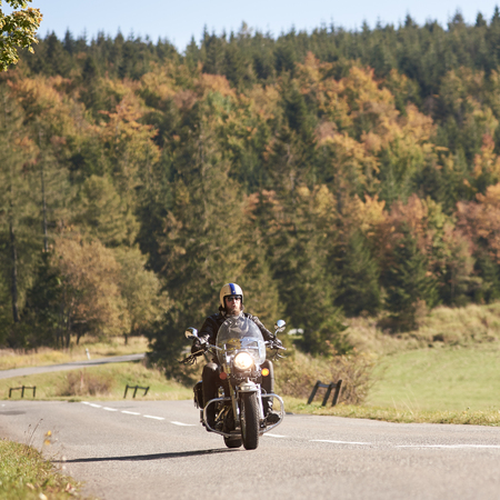 Bearded motorcyclist in helmet, sunglasses and black leather clothing riding cruiser motorbike along empty asphalt road on bright sunny autumn day.の写真素材