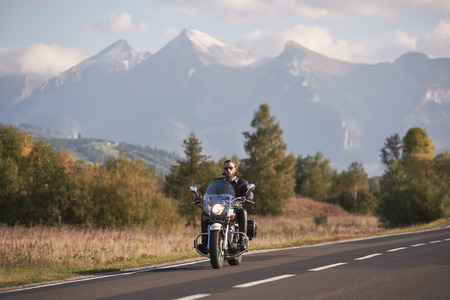 Bearded biker in sunglasses and black leather clothing riding modern powerful cruiser motorbike on blue foggy mountains with white snowy peaks and bright autumn sky copy space background.の写真素材