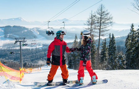 Pair of snowboarders on top of a ski slope at winter resort downhill on a sunny day. Man holds a womans hand against the backdrop of a ski-lift and snow-capped mountains. Bukovelの写真素材