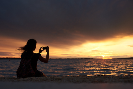 Dark silhouette of tourist woman with backpack sitting alone on seashore at water edge, taking picture of beautiful seascape at sunset on dark evening sky background. Tourism and vacations concept.の写真素材