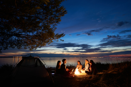 Night summer camping on sea shore. Group of young tourists sitting, laughing in high grass around bonfire near tent under beautiful blue evening sky. Tourism, friendship and beauty of nature concept.の写真素材