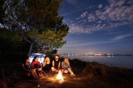 Group of five travellers, young man and women sitting on sea shore at campfire under tree. Tourist tent, quiet water surface and blue evening sky background. Tourism, friendship and camping concept.の写真素材
