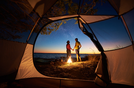View from inside tourist tent. Couple, man and woman hikers standing at campfire holding hands on lake shore on evening starry sky at sunset and blue lake water background. Tourism and love concept.の写真素材