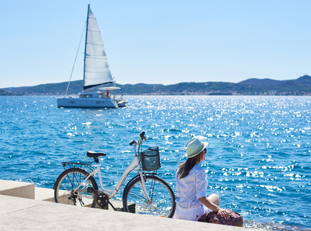 Perfect summer landscape on bright sunny day. Back view of female tourist with backpack sitting at bicycle on paved sidewalk under clear blue sky. Sailing ship in azure water on backgroundの写真素材
