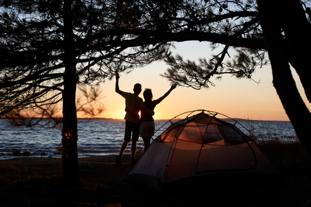 Back view of silhouettes of young tourist couple man and woman standing embraced on sea shore in front of small tent on blue clear water background enjoying quiet evening. Tourism and camping concept.の写真素材