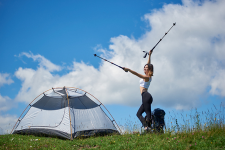 Happy slim woman tourist with backpack near tent, on the top of a hill against blue sky and clouds, lifting hands up in the air with trekking sticks in hands pointing into distance, in the mountainsの写真素材