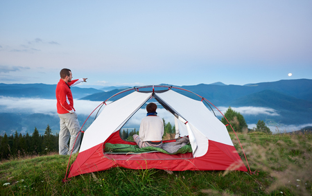 Young tourist male shows away to the moon his companion female. The girl sits in a tent near which there is a guy on the background of the beautiful scenery of the morning mountains at dawnの写真素材