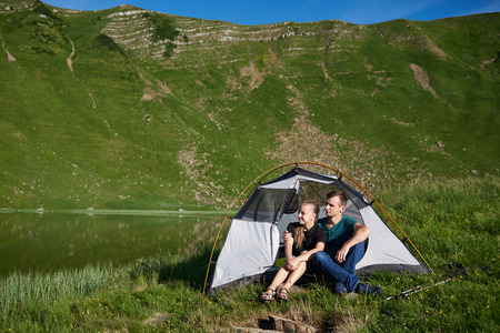 Young people sit in a tent and look out into the distance near a mountain lake against the backdrop of a green mountain on a sunny summer day.の写真素材