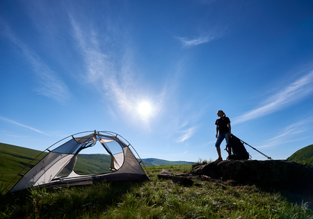 Young happy woman tourist with backpack and trekking sticks near tent, standing on big stone on the top of a hill against blue sky, sun and clouds, enjoying summer day in the mountainsの写真素材