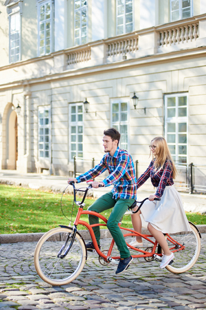 Active romantic tourist pair, bearded man and smiling long-haired blond woman in white skirt riding tandem bike along paved city sidewalk on bright sunny day on blurred ancient building background.の写真素材