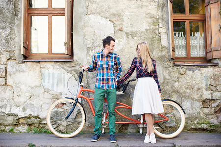 Young attractive romantic tourist couple, bearded man and blond woman looking to each other, standing together at modern tandem bicycle on empty pavement at cracked wall of old residential house.の写真素材