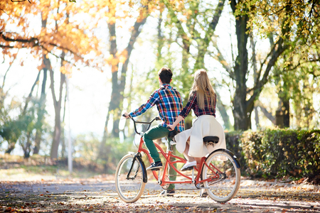 Back view of young tourist couple, man and blond long-haired woman cycling together tandem double red bicycle by park or forest sunny alley covered with golden leaves at autumnの写真素材
