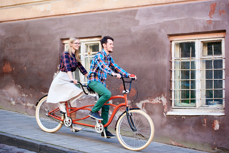 Young handsome man and pretty blond woman riding together tandem double bicycle along paved sidewalk on bright sunny autumn day by old buildings with cracked walls and iron lattice on windows.の写真素材