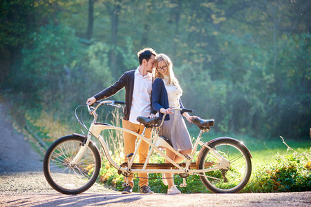 Young happy romantic couple, bearded man and attractive woman close together at tandem double bicycle outdoors in summer park or forest on blurred sunny green trees dense foliage background.の写真素材