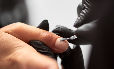 Process of making manicure in salon. Close-up of manicure master hand in black rubber gloves applying with brush transparent gel polish on woman fingernails on blurred background. Fashion and beauty.の写真素材