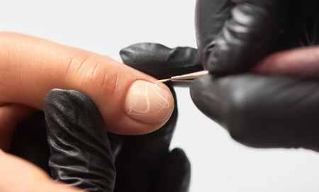 Fashionable manicure in beauty salon. Close-up of professional manicurist hands in black rubber gloves painting elegant abstract patterns on client woman nails using applicator on white background.の写真素材