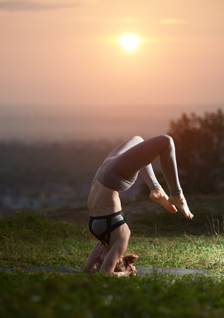 Sportive muscular flexible girl in difficult balancing yoga posture outdoors on green grassy meadow on copy space background of clear light pink summer sky. Fitness, training and beauty concept.の写真素材