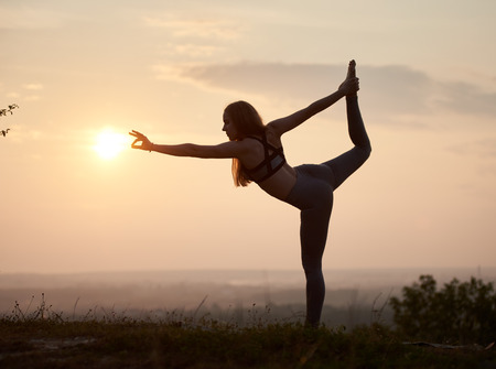 Silhouette of slim strong muscular girl standing on one leg doing complicated yoga exercises outdoors at dawn on copy space background of light pink summer sky and blurred misty horizon. Narajasanaの写真素材