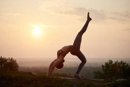 Silhouette of slim strong muscular girl with raised leg doing complicated yoga exercises on grassy hill on copy space background of bright light summer sky at sunset.の写真素材
