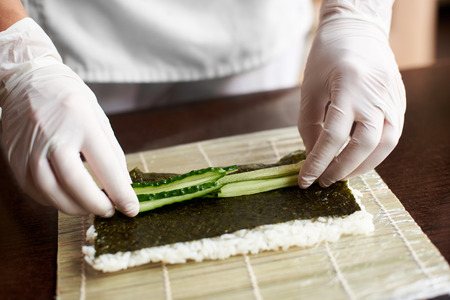 Close-up view of process of preparing rolling sushi. Nori, white rice and cucumber on bamboo mat. Chefs hands in gloves starts cooking sushi rollsの写真素材