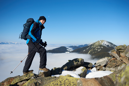 Tourist hiker with backpack and trekking poles climbing up on steep rocky mountain slope on background of blue sky, foggy valley filled with white clouds and snowy mountain tops.の写真素材
