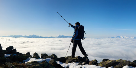 Tourist hiker with backpack standing on huge rocks pointing with trekking pole at bright blue sky on background of foggy valley filled with white clouds and distant mountain tops.の写真素材
