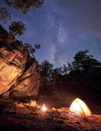Mountain campsite at summer evening amid huge steep rock formation. Small tourist tent brightly lit by burning campfire under clear dark starry sky. Tourism, climbing, hiking and traveling concept.の写真素材