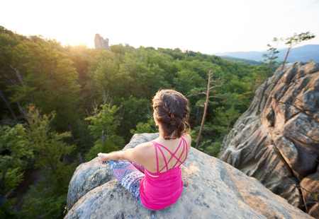 Aerial view of lit by summer sun young slim sporty woman sitting on top of huge rock, doing yoga on green trees tops forest background in the morning. Sport, gymnastics and fitness concept. Siddhasanaの写真素材
