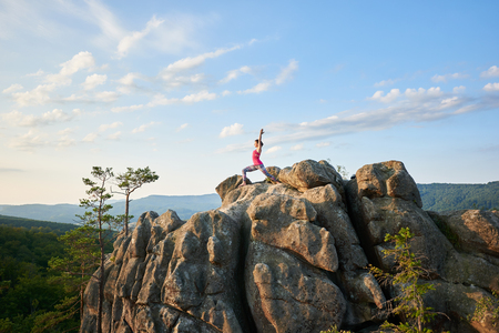 Young slim woman standing with raised hands in yoga pose on top of rocky mountain in the summer morning, clear blue sky on background. Sport, gymnastics and healthy lifestyle concept. Virabhadrasanaの写真素材