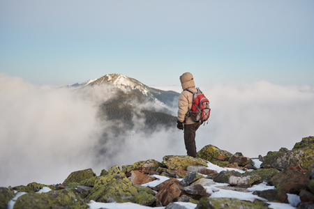 Back view of tourist traveler in warm clothing with backpack on the top of rocky mountain enjoying view of low white clouds, snowy woody mountain peak and bright blue morning sky at sunrise.の写真素材
