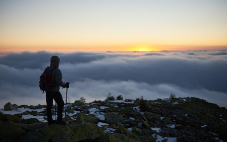 Back view of tourist in warm jacket with backpack and hiking sticks on rocky mountain peak on background of valley covered with white clouds stretching to horizon and bright morning sky at down.の写真素材